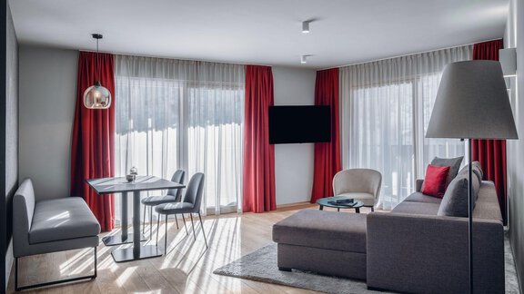 A modern living room with a cozy couch and a dining table. Large windows let in plenty of light, and the red curtains provide a striking accent. - AlpenParks The Secret Sölden - Sölden