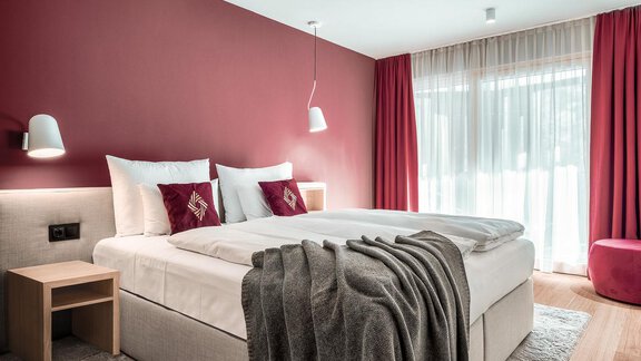 A modern bedroom with a cozy double bed and red accents. The windows are dressed with light curtains that let in plenty of light. - AlpenParks The Secret Sölden - Sölden