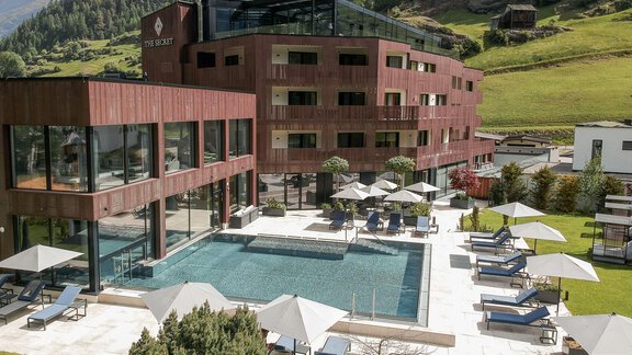 A modern hotel with a large swimming pool and sun loungers in the garden. In the background, green mountains can be seen. - AlpenParks The Secret Sölden - Sölden
