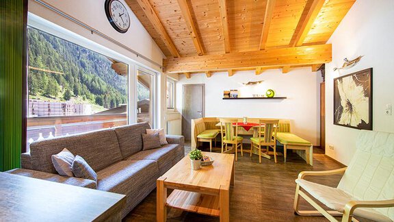 A modern living room with wooden ceilings and large windows. The space is cozily furnished with a sofa, a table, and colorful chairs. - Alpensweet - Sölden