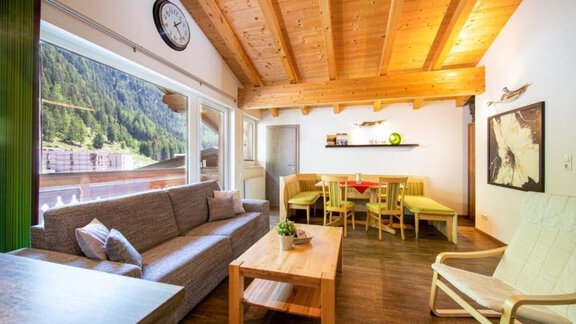 A cozy living space with a wooden beam ceiling and a large window overlooking the mountains. There is a sofa, a wooden table, and colorful chairs. - Alpensweet - Sölden