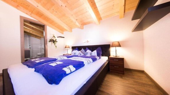 A cozy bedroom with a double bed and wooden beam ceiling. The blue bedspread and a bedside lamp give the room an inviting atmosphere. - Alpensweet - Sölden