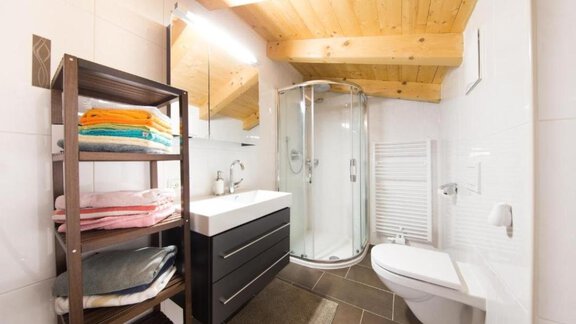 A modern bathroom with a shower area and wooden beams on the ceiling. Colored towels are lying on a shelf in the corner. - Alpensweet - Sölden