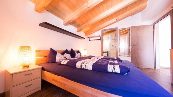 A modern bedroom with a large bed and wooden ceilings. There is a bedside lamp and access to another room. - Alpensweet - Sölden