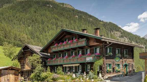 A traditional wooden house in the mountains with colorful flowers. The surroundings are green and wooded, under a bright sky. - Altes Holzhaus - Sölden