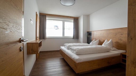 A simple bedroom with a double bed and two pillows. The room has a wooden floor and a large window that lets natural light in. - Anraitl Appartements Sölden - Sölden