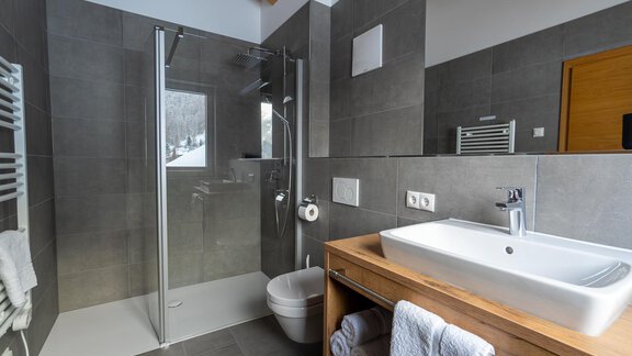 A modern bathroom with gray tiles and a separate shower. The amenities include a sink on a wooden cabinet and towels. - Anraitl Appartements Sölden - Sölden