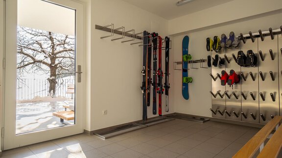 A spacious ski room with skis and snowboards on the walls. Through the window, you can see a snowy outdoor area with a tree. - Anraitl Appartements Sölden - Sölden