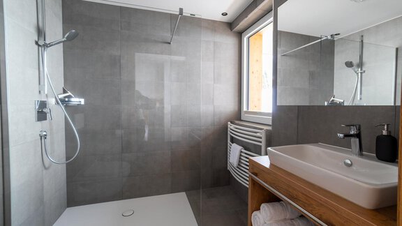 A modern shower with gray tiles and a large mirror. Fresh towels are ready on the wooden shelf. - Anraitl Appartements Sölden - Sölden