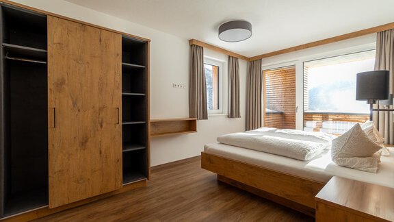 A modern bedroom with wooden furniture and a large window. There is a bed, a wardrobe, and a desk. - Anraitl Appartements Sölden - Sölden