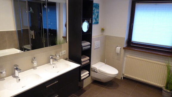 Ein modernes Badezimmer mit einem Doppelwaschtisch und großen Spiegeln. Der Raum ist hell und ordentlich, mit einer Toilette und Handtüchern in einem Regal. - Apart 55 - Sölden