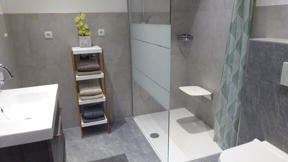 Ein modernes Badezimmer mit einer Dusche aus Glas und grauen Fliesen. Auf der linken Seite befindet sich ein Waschbecken und ein Regal mit Handtüchern. - Apart 55 - Sölden