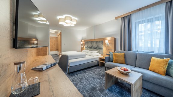 A modern hotel room with a comfortable bed and a cozy sofa. The room is bright and friendly with wooden details and a television. - Apart Hotel Garni Alt Kaisers - Sölden