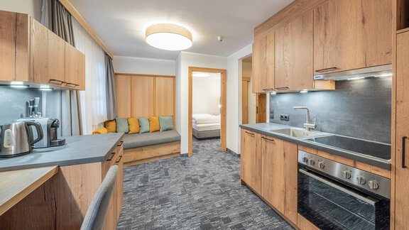 A modern kitchen unit with wooden cabinets and a dining area. In the background, a cozy living area with a sofa can be seen. - Apart Hotel Garni Alt Kaisers - Sölden