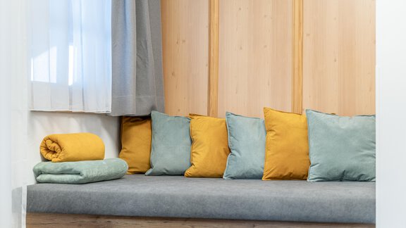 A cozy seating area with a gray bench and colorful cushions in yellow and green. In the background, bright curtains and wooden panels can be seen. - Apart Hotel Garni Alt Kaisers - Sölden