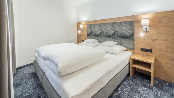 A modern bedroom with a large double bed and stylish wooden paneling. The walls are light, and there is a bedside lamp next to the bed. - Apart Hotel Garni Alt Kaisers - Sölden
