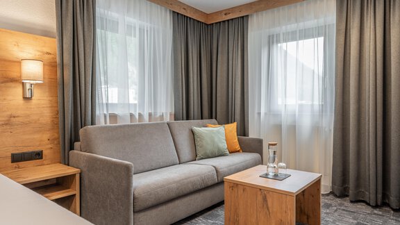 A cozy room with a gray sofa and a wooden table. Large windows with curtains let in plenty of light. - Apart Hotel Garni Alt Kaisers - Sölden