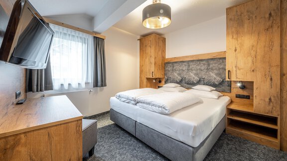 A modern bedroom with a large double bed and wooden furniture. The room is bright and inviting with a window and a television. - Apart Hotel Garni Alt Kaisers - Sölden