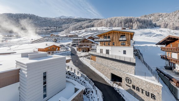 A snow-covered landscape with modern buildings and traditional chalets. In the background, snowy mountains and a blue sky can be seen. - Apart Natur - Sölden