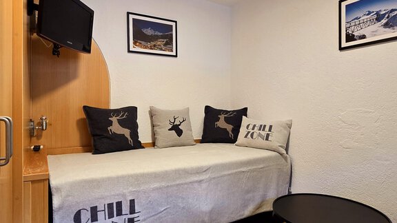 A cozy room with a single bed, pillows, and a television. The walls are decorated with pictures, and the floor is made of wood. - Apart Tyrolis - Sölden
