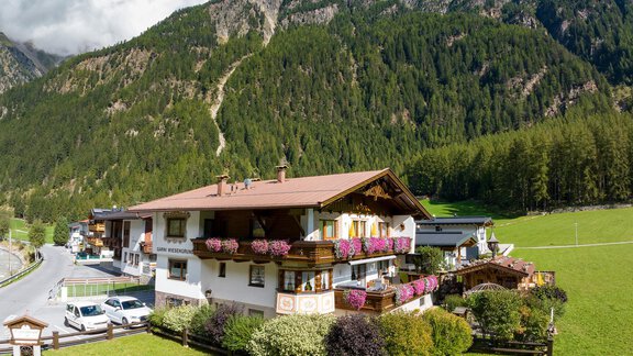 A charming house in the mountains with blooming plants and a beautiful view of nature. In the background, there are wooded hills and a blue sky. - Apart Wiesengrund - Sölden