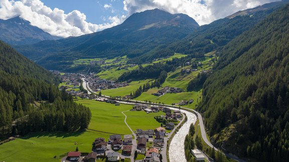 A picturesque mountain landscape with gently rolling hills and a clear river. In the foreground lies a small village, surrounded by green fields and forests. - Apart Wiesengrund - Sölden