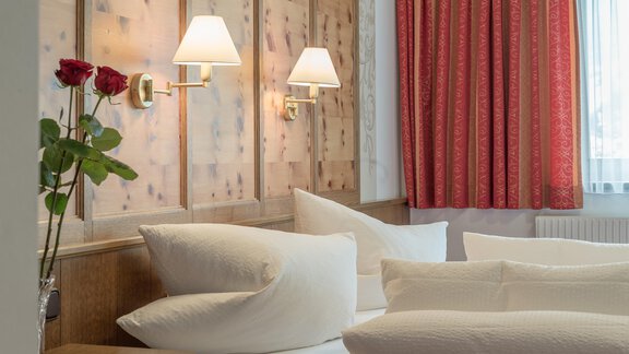 A cozy bedroom with a wood panel and warm light. A rose is on the nightstand, and the windows are decorated with red curtains. - Apart Wiesengrund - Sölden