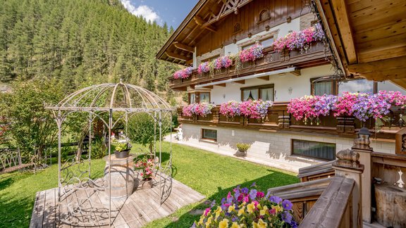 A charming wooden house with flower balconies and a beautiful garden landscape. In the foreground stands a stylish pergola surrounded by colorful flowers. - Apart Wiesengrund - Sölden