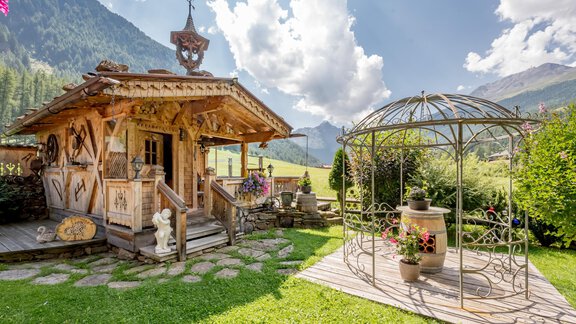 A picturesque wooden cabin in a green landscape with mountains in the background. Next to the cabin stands an elegant garden gazebo with flowers. - Apart Wiesengrund - Sölden