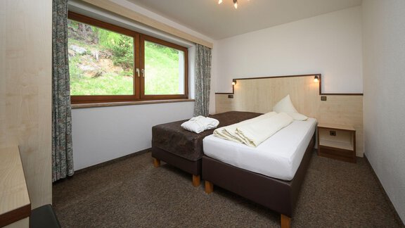 A simple hotel room with two beds and a window that looks out onto a green landscape. The walls are light and there is a small table in the room. - Appartement Alpin - Sölden