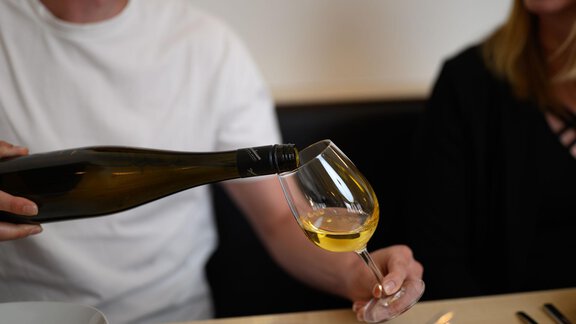 A person is pouring wine into a glass. In the background, another person can be seen enjoying the moment. - Appartement Alpin - Sölden