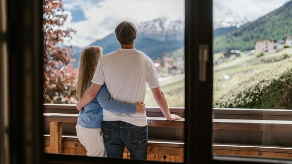 A couple is standing on a balcony, looking out at a beautiful mountain landscape. In the background, there are snow-covered mountains and green meadows. - Appartement Alpin - Sölden