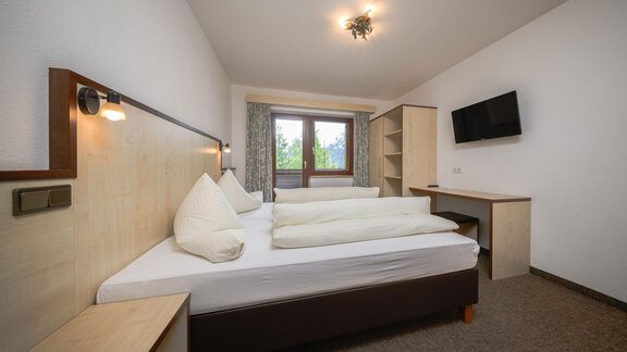 A modern bedroom with a double bed and two pillows. The room has a desk, a television, and large windows with a view outside. - Appartement Alpin - Sölden