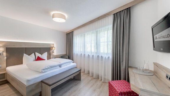 A modern bedroom with a large bed and fresh white bedding. The room has bright walls and a large window with curtains. - Appartement Cristallo - Sölden