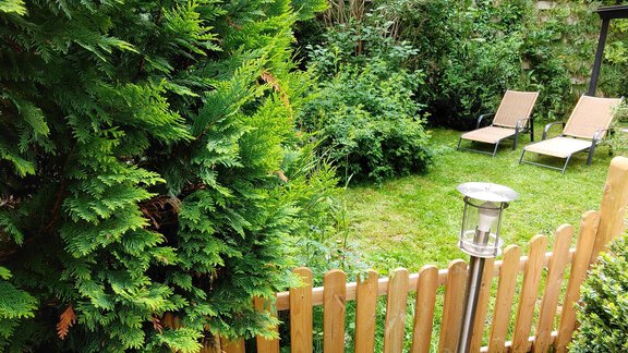 A quiet garden with lush greenery and a wooden slatted fence. Two loungers are ready for relaxing. - Appartement Gehörde Sölden - Sölden