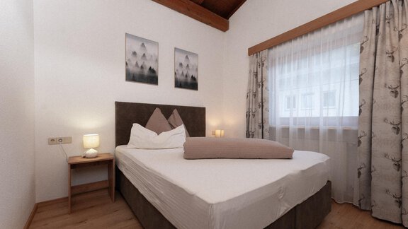 A cozy bedroom with a large bed and wooden ceilings. Soft colors and a window with curtains create an inviting atmosphere. - Appartement Gehörde Sölden - Sölden