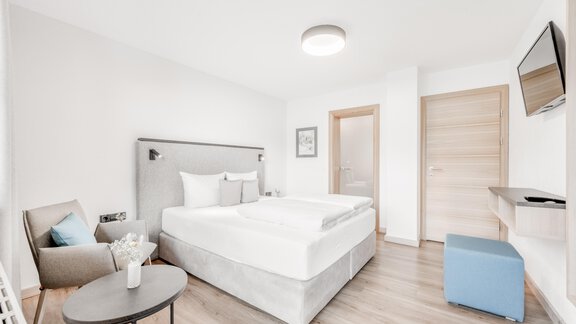 A modern bedroom with a large bed and minimalist furnishings. There is an armchair, a table, and a television on the wall. - Appartement Montanara - Sölden