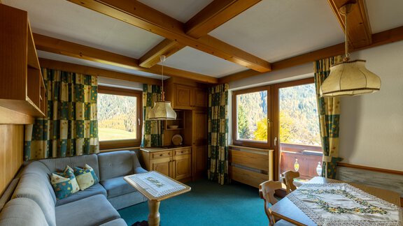 A cozy living room with a wooden ceiling and sunny outlook. The furnishings consist of a sofa, a table, and decorative curtains. - Appartementhaus Karlinger - Sölden