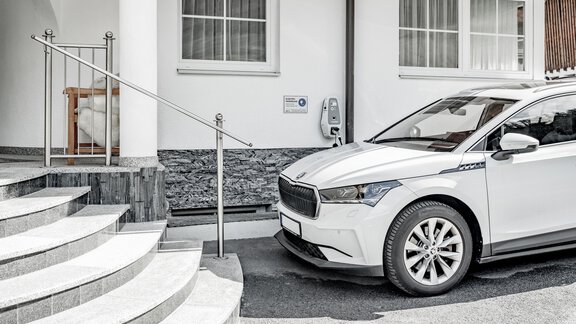 A modern, white car is parked in front of a building. In the background, stairs and an area for charging electric vehicles are visible. - Appartementhaus Kathrin - Sölden