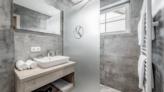 A modern bathroom with gray walls and a transparent shower. Fresh towels are placed on the wooden shelf and a sink is installed. - Appartementhaus Kathrin - Sölden