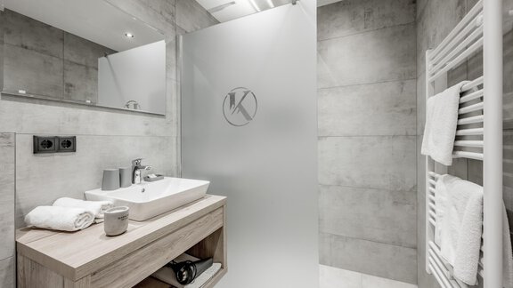 A modern bathroom with gray walls and a transparent shower partition. A wooden console with a sink and towels complements the decor. - Appartementhaus Kathrin - Sölden