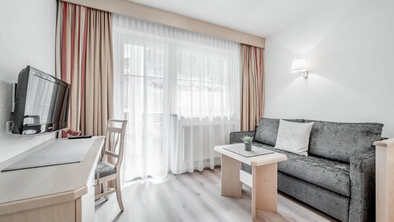 A bright living room with a gray sofa and a table. There is a television and large windows with curtains. - Appartementhaus Kathrin - Sölden