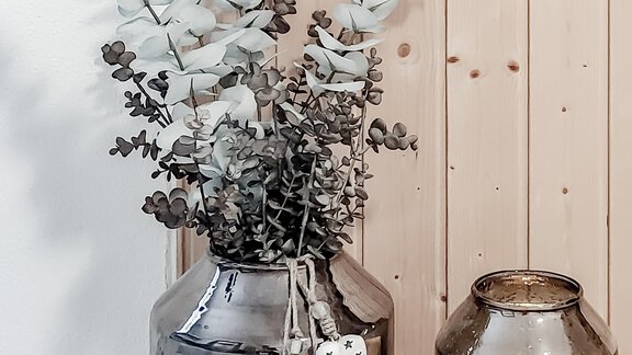 A modern vase with eucalyptus branches stands next to another elegant vase. The background is made of light wooden boards, creating a cozy atmosphere. - Appartementhaus Kathrin - Sölden