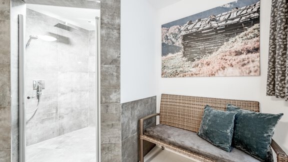 A modern bathroom with a glass shower and gray tiles. Next to the shower is a cozy bench with cushions and a decorative picture on the wall. - Appartementhaus Kathrin - Sölden