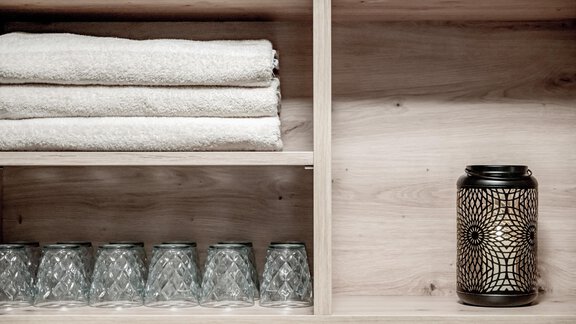 A decorative shelf with stacked towels and glass glasses. On the right side, there is a black lantern with a unique pattern. - Appartementhaus Kathrin - Sölden