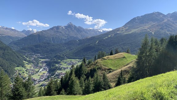 A green, hilly landscape with high mountains in the background. The sky is clear and blue, and the village is situated in a valley. - Appt. Ideal - Sölden