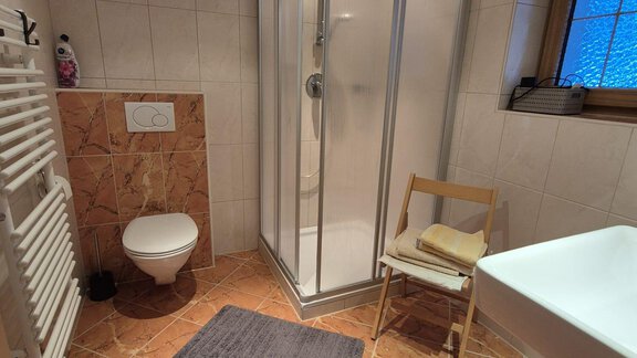 A modern bathroom with a shower, a toilet, and a sink. The floor is tiled, and there is a chair as well as a towel holder. - Appt. Wildschütz - Sölden