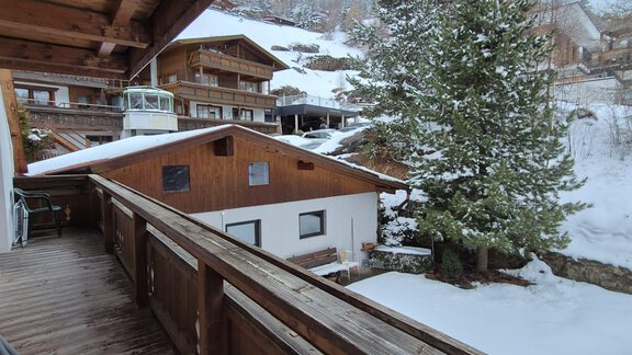 A view from a wooden staircase overlooking a snowy winter village. In the foreground, wooden houses and a green tree are visible. - Appt. Wildschütz - Sölden