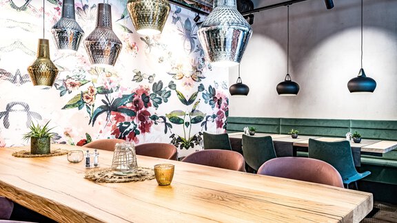 Ein modern eingerichteter Raum mit einem großen Holztisch und bunten Wandleuchten. Die Wände sind mit floralen Muster dekoriert, und es gibt grüne Sitze für die Gäste. - Bäckelar Wirt - Sölden