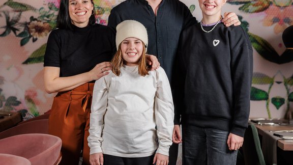 Eine Gruppe von vier Personen posiert gemeinsam vor einer bunten Blumenwand. Sie stehen in einem modernen Café oder Restaurant. - Bäckelar Wirt - Sölden